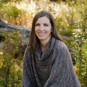 Tara Vicars sits on a log outside, smiling, dressed in a shawl and jeans with autumn colors behind her.